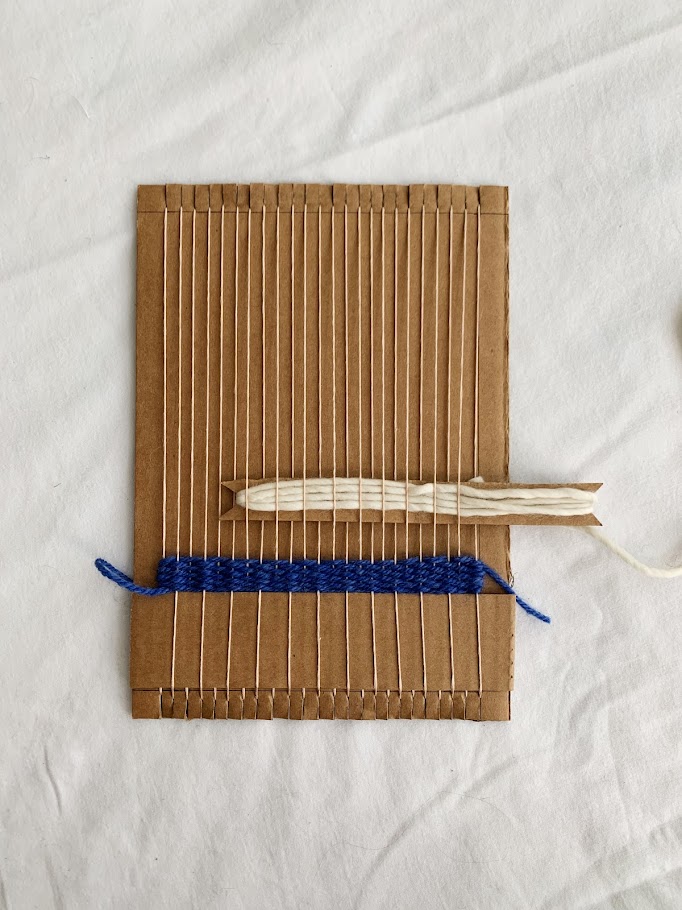A cardboard shuttle with cream colored yarn that is partially inserted into a cardboard loom
