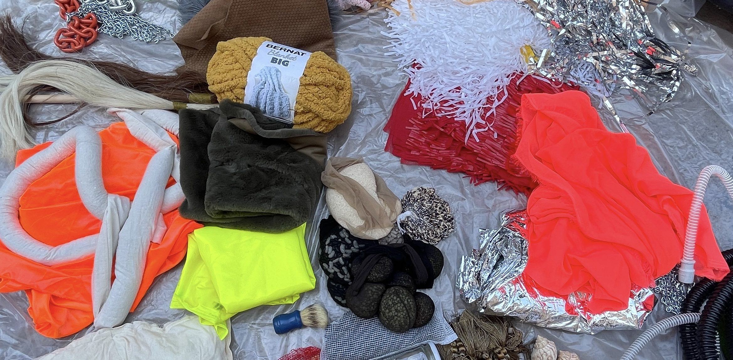 [objects on ground] A wide array of objects arranged haphazardly on a sheet of clear plastic. Reflective silver mylar pom poms, neon orange fabric and red latex gloves, interspersed with tan and white conch shells, beans in black tights, brown faux fur, gold, pink and magenta thick intestine-like yarns and tan and brown horsehair.