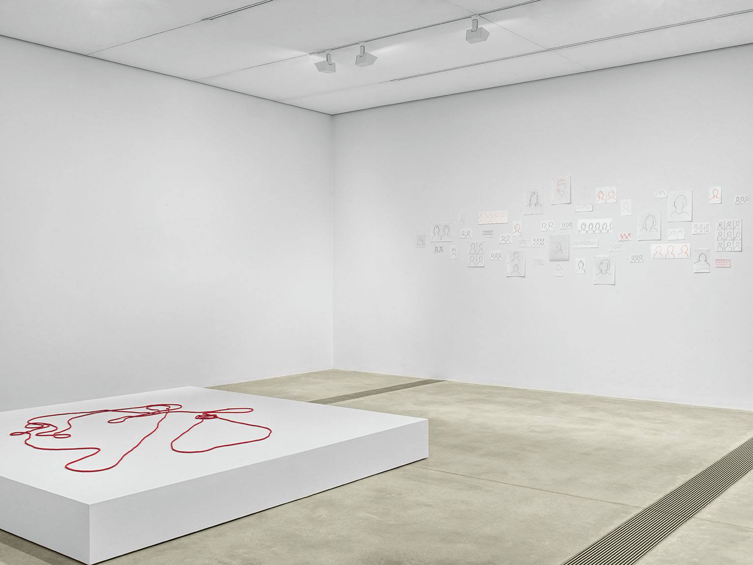 Red beaded rope displayed on white platform with several silhouette drawings posted on the wall to the right