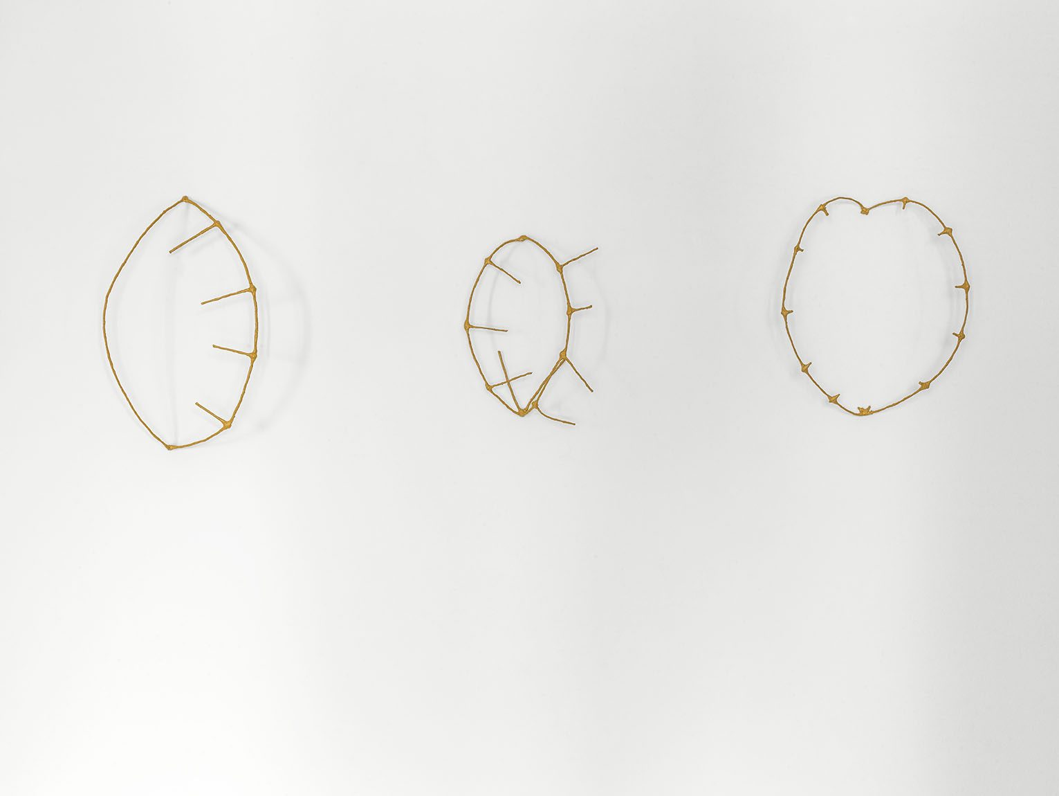 Three yellow wire sculptures in the shape of ochre traps hanging side by side on a white wall