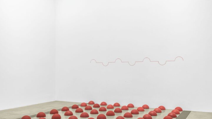 Several small red sculptures in the shape of little mounds laying on the concrete ground in a grid form with a red line drawn across the wall with several peaks