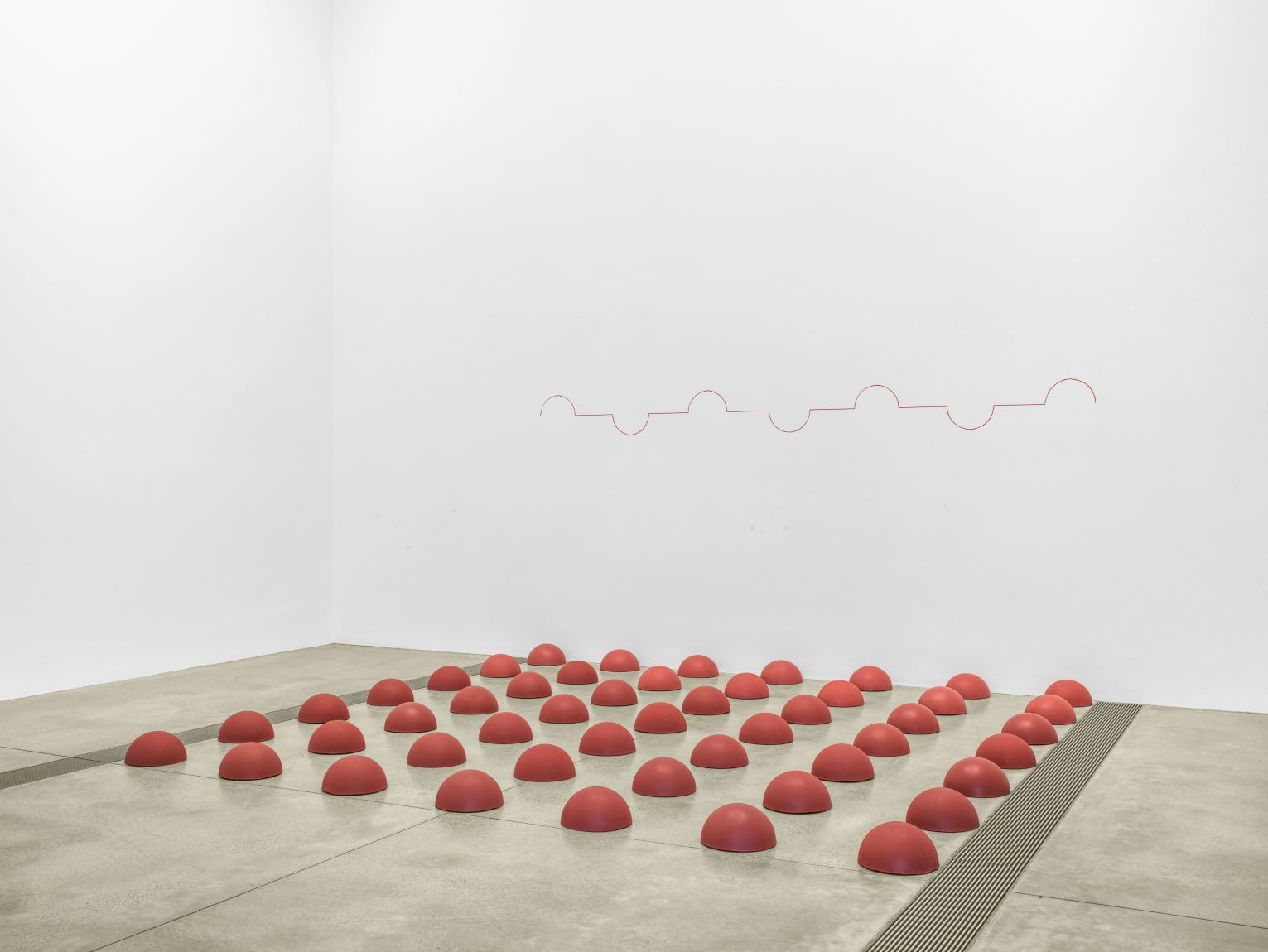 Several small red sculptures in the shape of little mounds laying on the concrete ground in a grid form with a red line drawn across the wall with several peaks