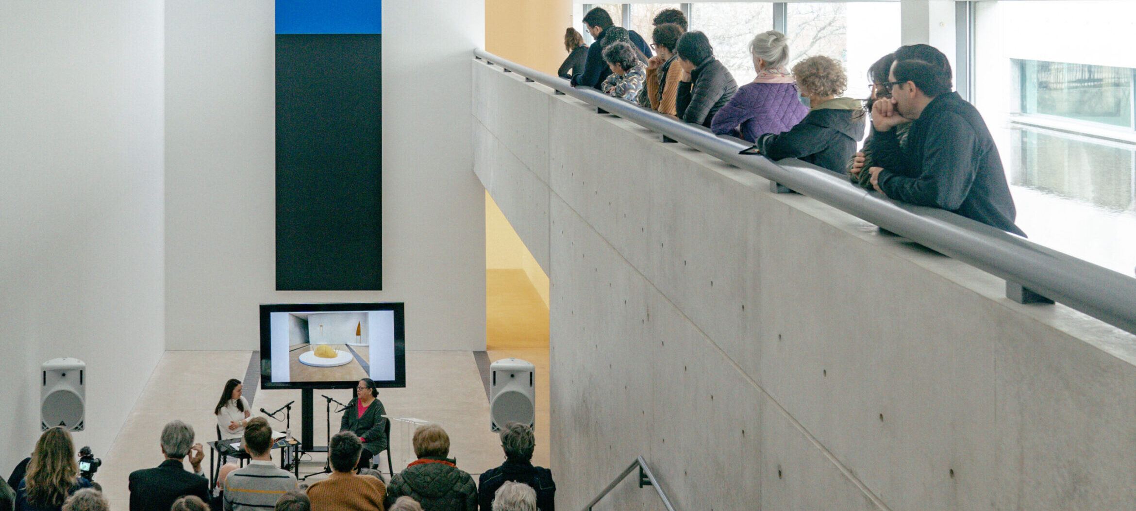 Artist Talk program with Faye HeavyShield and curator Tamara Schenkenberg, sitting in front of blue and black vertical painting, surrounded by an audience