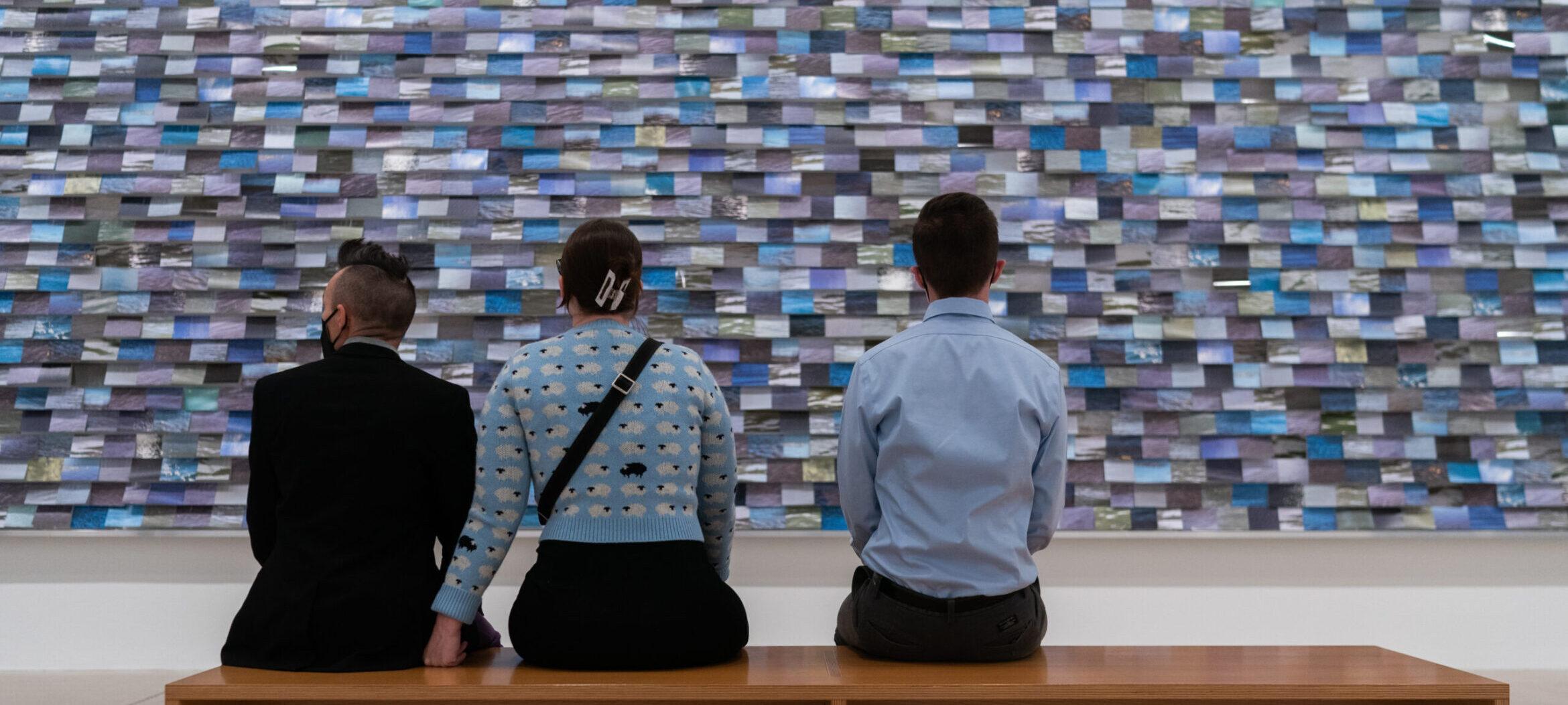Three visitors on a bench looking at Faye HeavyShield commission, "aiyo niitahtaan." A large blue installation with thousands of 4x6 images of rivers.