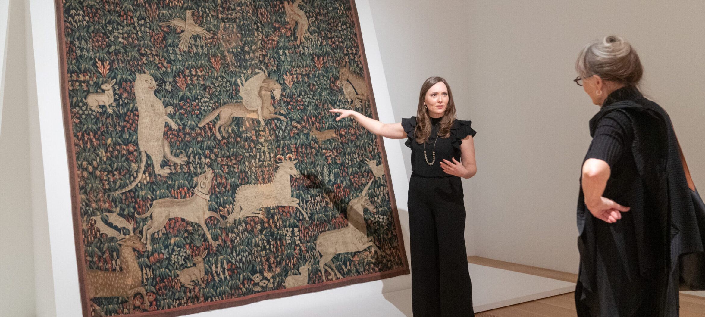 Assistant Curator, Heather Alexis Smith, point to the "Millefleurs Tapestry," a large floral tapestry with several animals and mythical creatures, hung at an angle on a white plinth.