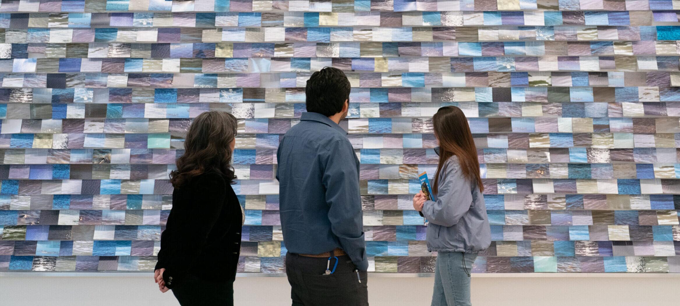 Three individuals looking at a large horizontal installation with thousands of 4x6 blue images of rivers.