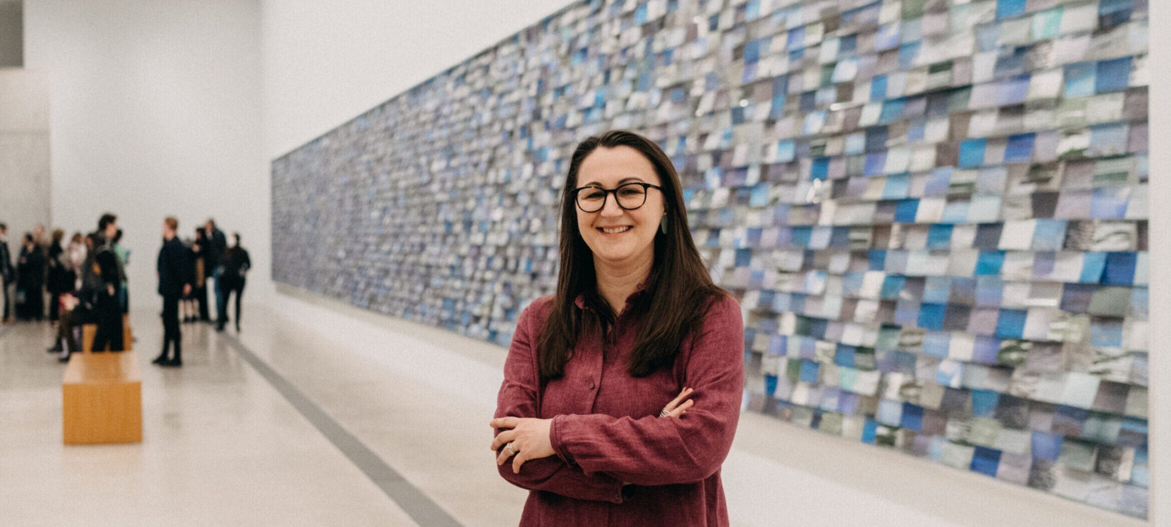 Pulitzer curator Tamara Schenkenberg wearing a red button down shirt, posing with arms crossed in front of large horizontal installation of thousands of 4x6 photographs of rivers.