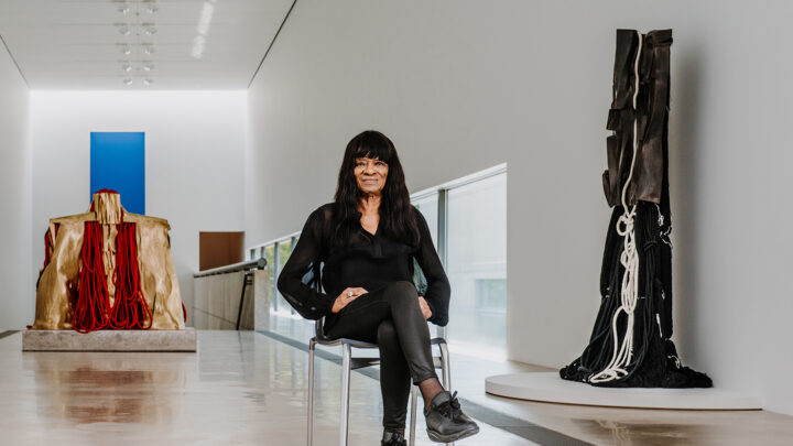 Artist Barbara Chase-Riboud wearing black leather pants and a longe sleeve sheer black blouse, sitting in front of her bronze sculpture with bright red rope