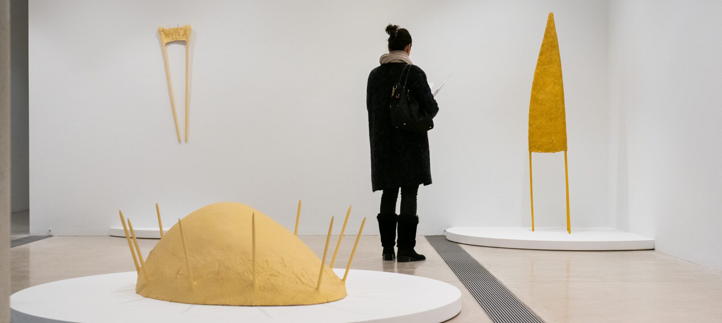 Person observing a 3-sided yellow paper mached sculpture, with a sculpture on the ground with spikes in the hair and another sculpture shaped like a large wishbone on the wall.