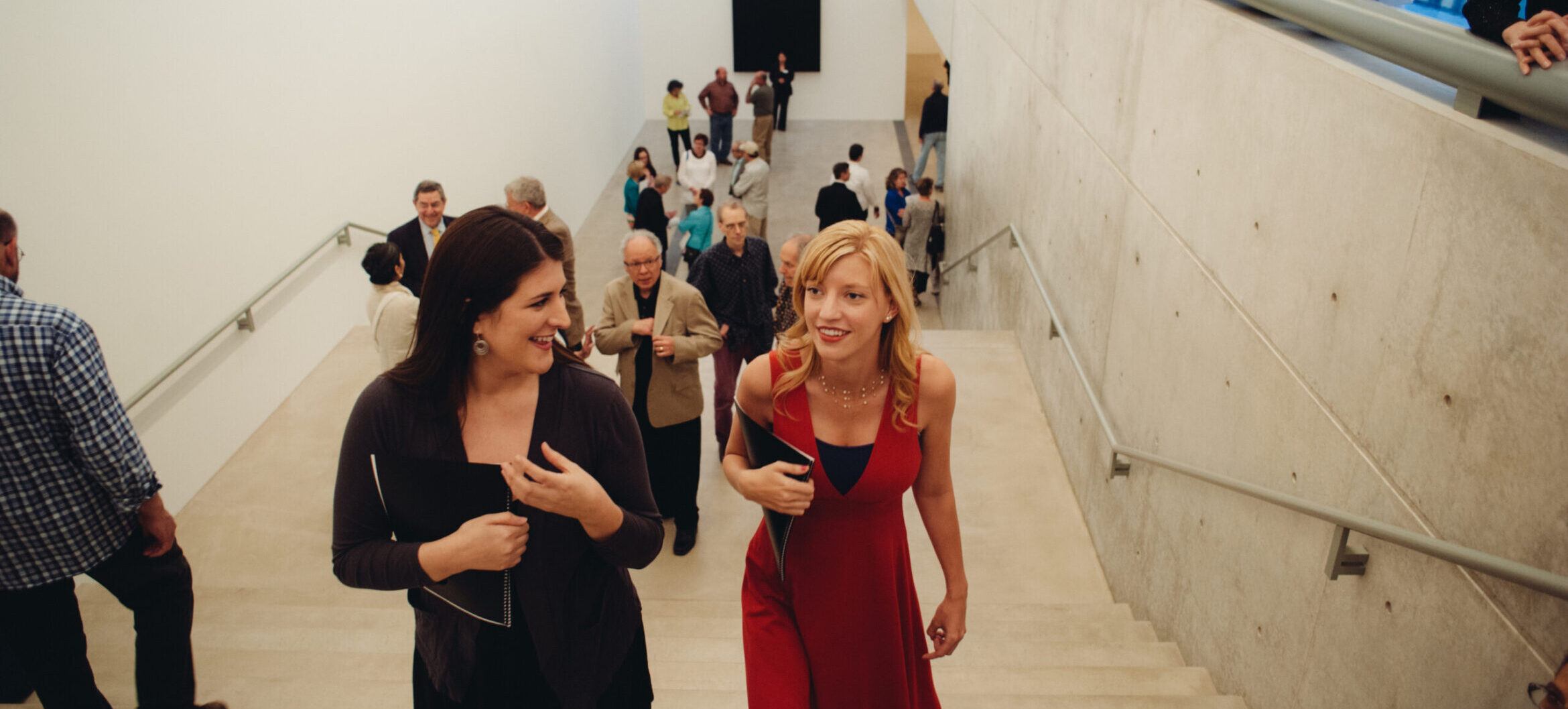 A visitor in with brown hair in a black dress and one with blonde hair in a red dress walking up the stairs at a Pulitzer opening.