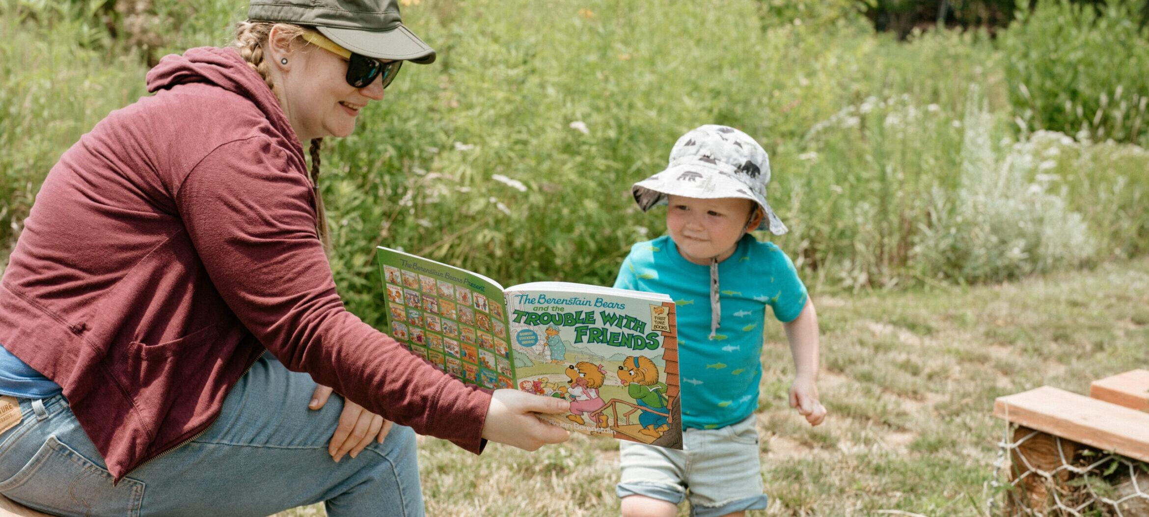Little boy in a bucket hat and aqua blue shirt, smiling and reading "The Berenstain Bears" with a Pulitzer staff member.
