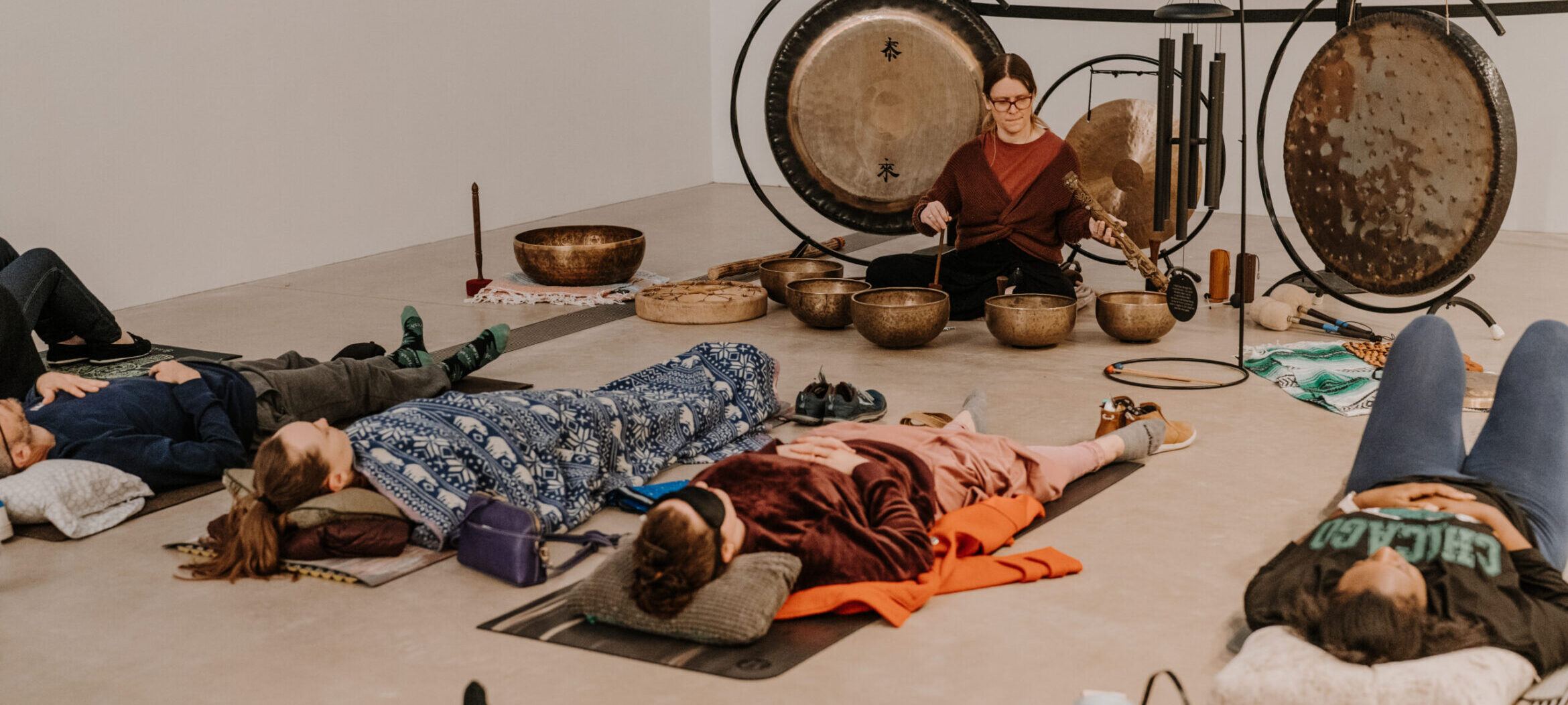 A group of people laying down in the gallery on yoga mats. Dr Aria sits in front surrounded by gongs and metal bowls