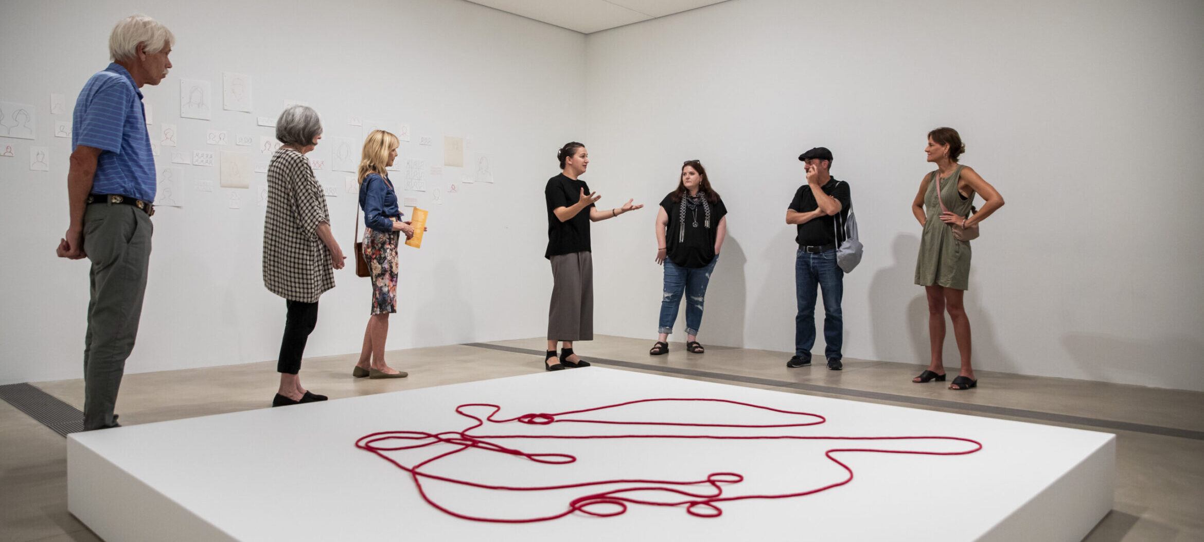 Visitors listening to a tour from Pulitzer Curator Tamara Schenkenberg, standing over a white platform with a bright red beaded rope by artist Faye HeavyShield