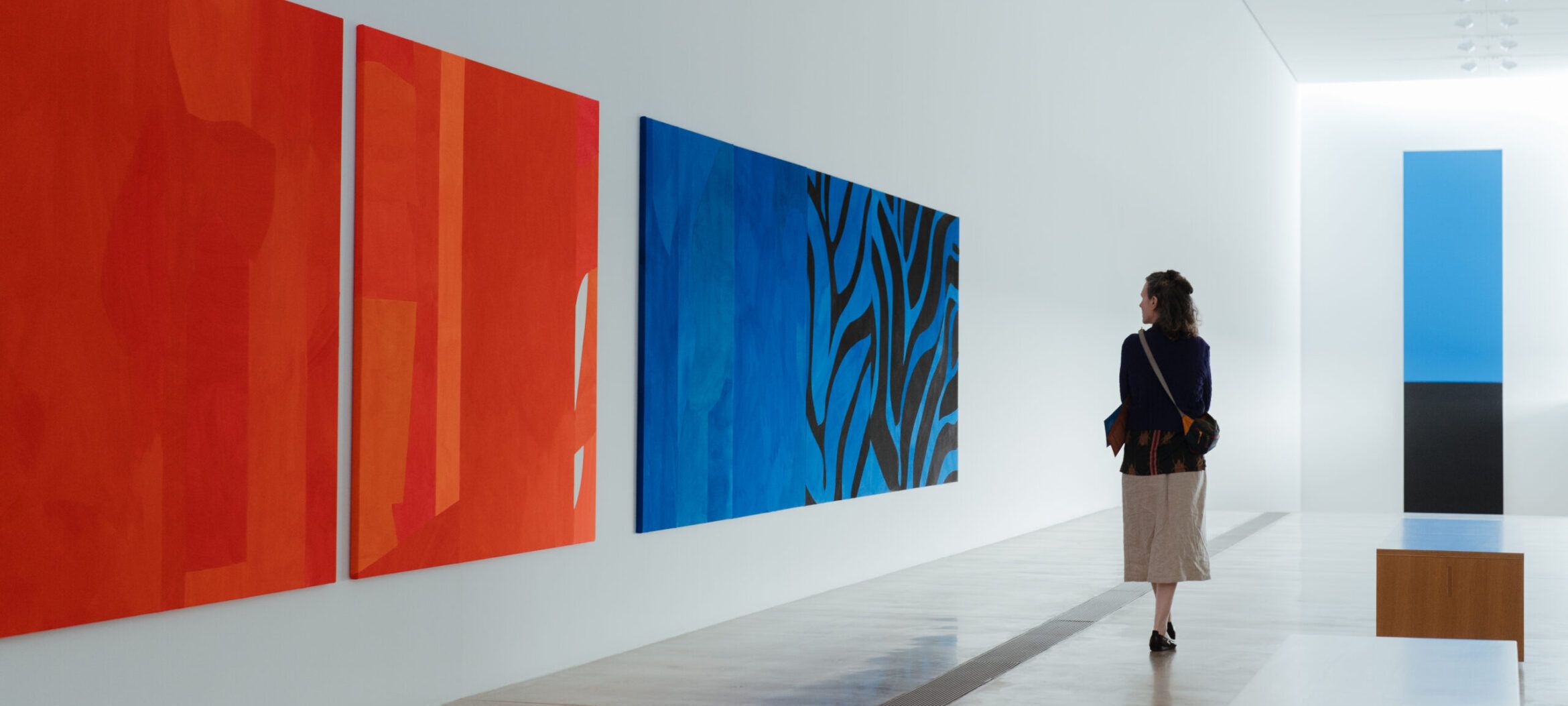 Woman looking at orange and blue Sarah Crowner painting with Ellsworth Kelly's Blue Black painting in the background.