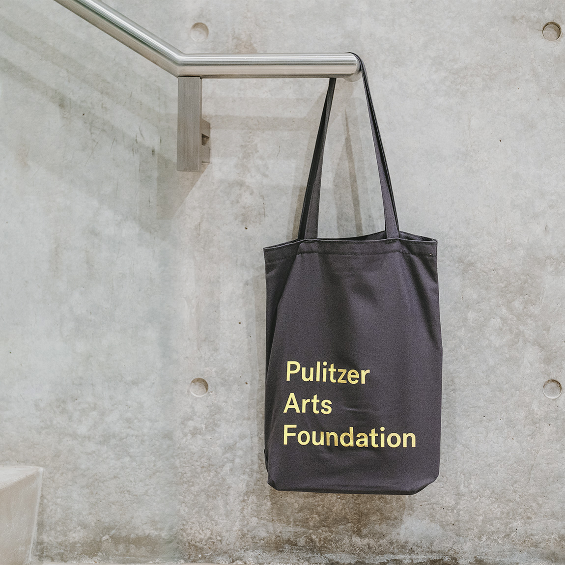 A grey tote bag hanging off a metal hand railing in front of a concrete wall