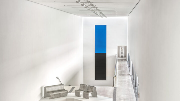 A modern art gallery with high white walls and a polished concrete floor, viewed from an elevated perspective. Several contemporary sculptures and furniture pieces are displayed on white platforms throughout the long, narrow space. A bright blue rectangular artwork hangs on the far wall, drawing the eye down the gallery.