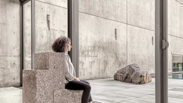 The image shows a modern, minimalist space with a person sitting on a geometric, stone-like bench that has a textured, almost granite-like surface. The individual is seated by large glass windows, facing outdoors where a smooth, concrete courtyard is visible, along with a large, natural rock. The room's interior walls are made of exposed concrete with a clean, industrial aesthetic. The serene atmosphere evokes a sense of contemplation, blending both natural and industrial elements.