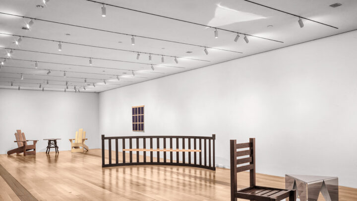 A spacious, minimalist gallery with light wood flooring and white walls. Several pieces of modern furniture are displayed on white platforms. These include a wooden chair with a slatted seat and back, a curved wooden railing, and two Adirondack chairs with a small table between them. A geometric, reflective sculpture sits on a platform in the foreground.