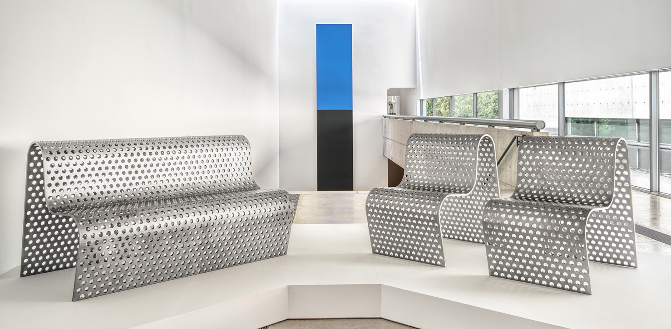  Modern metal furniture with a perforated design displayed on a white platform in a minimalist gallery space.