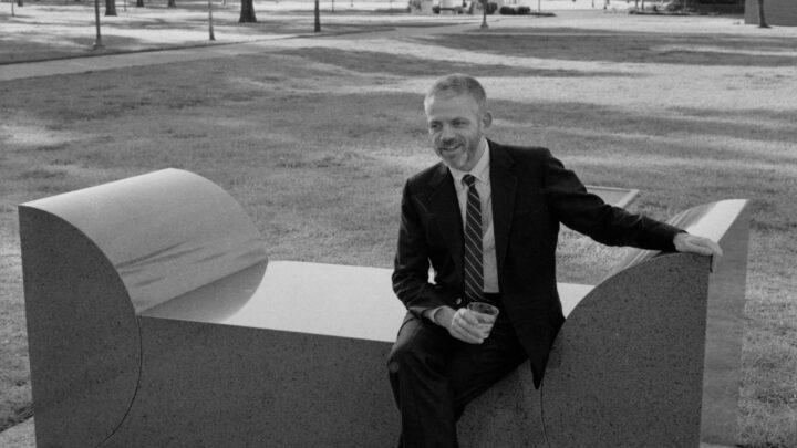 Man in a black suit holding a drinking cup sitting on an angled bench outdoors.