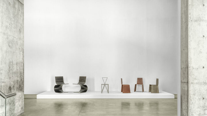 A minimalist gallery space with a polished concrete floor and off-white walls. Three sets of chairs are displayed on a low white platform. The first set includes two modern chairs with curved, ribbon-like backs, made of dark metal. The second set is a single geometric chair with thin, angular legs, made of a lighter metal. The third set consists of three simple chairs with blocky shapes and flat, reddish-brown seats and backs.