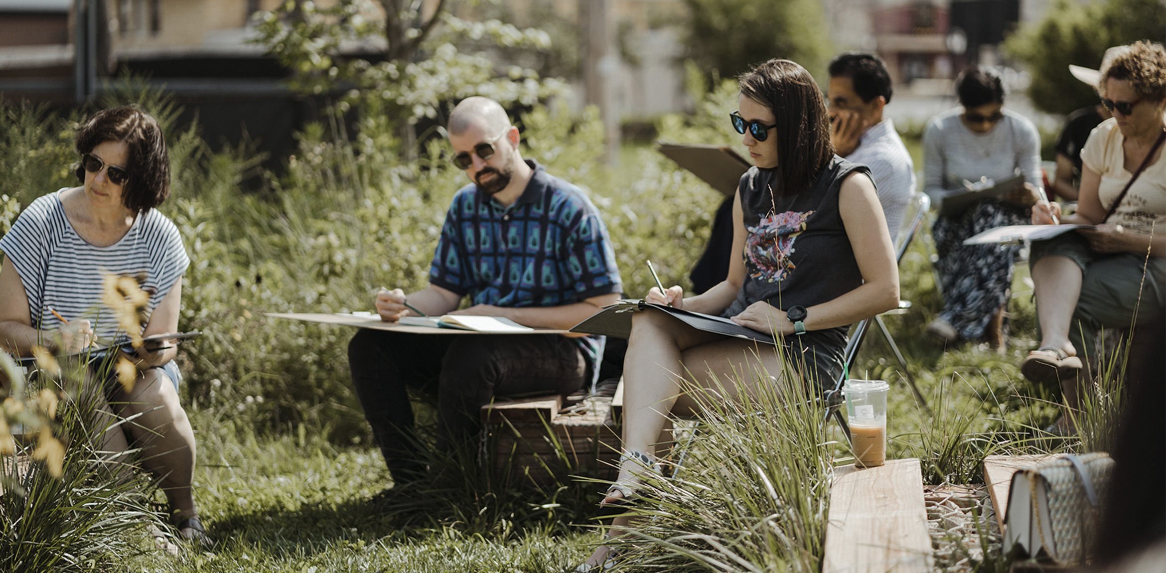 A group of people sitting on stools in a grassy park sketching and writing on large pads