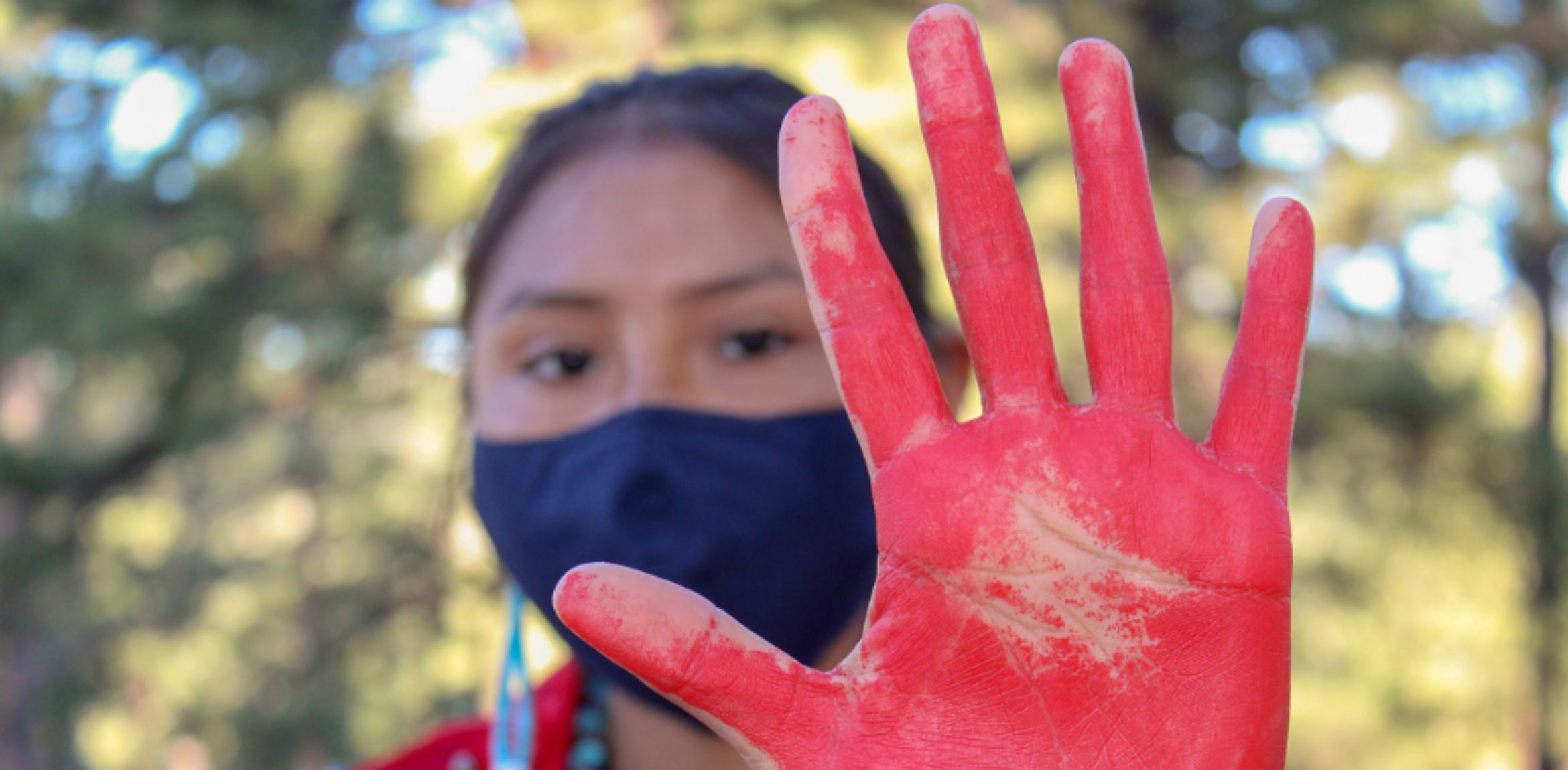 Indigenous woman with a black face mask holding hand up covered in red paint