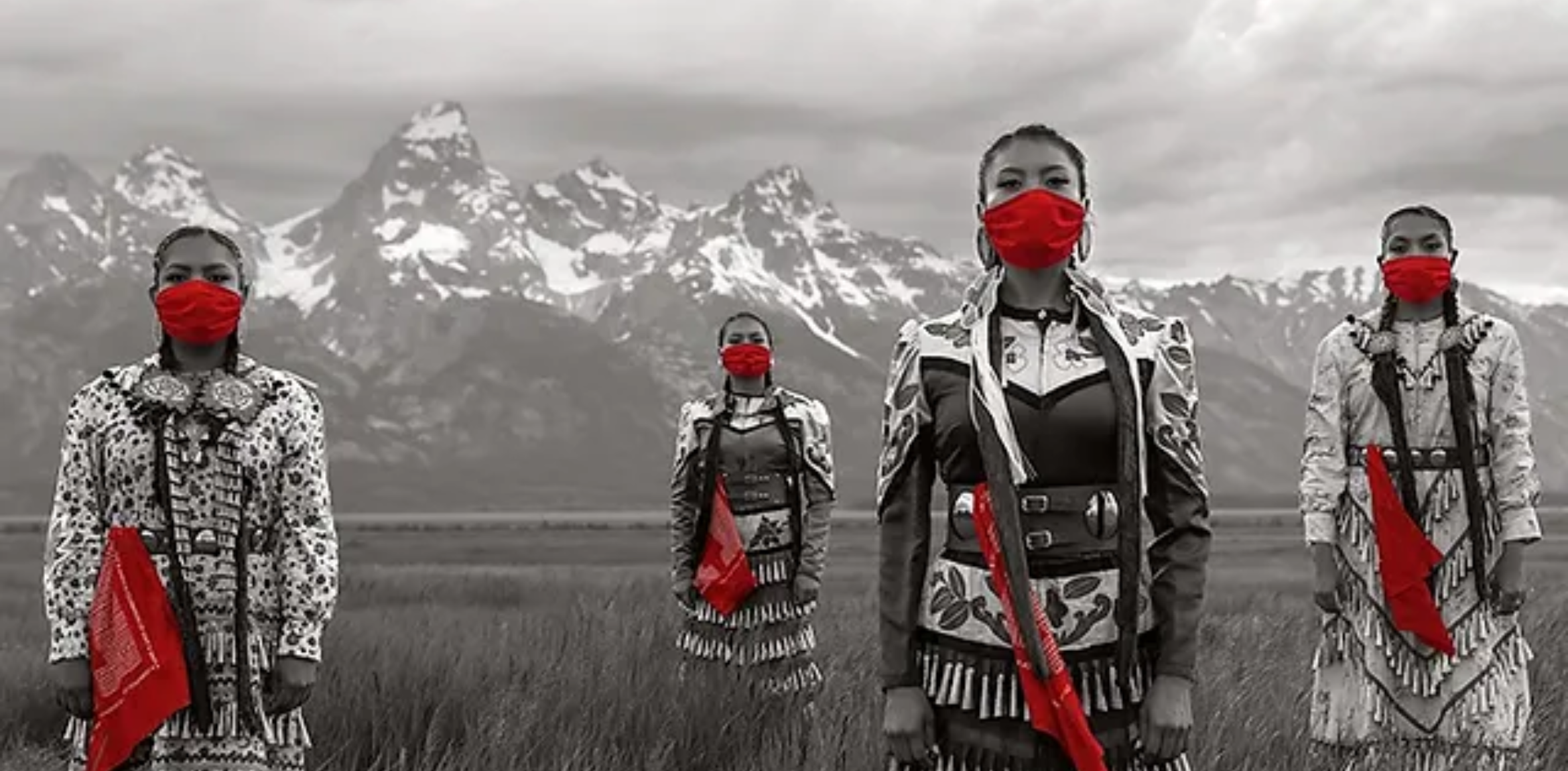 Black and white image of four indigenous woman with red face masks and red bandanas tied to their belts, standing in front of a mountain range