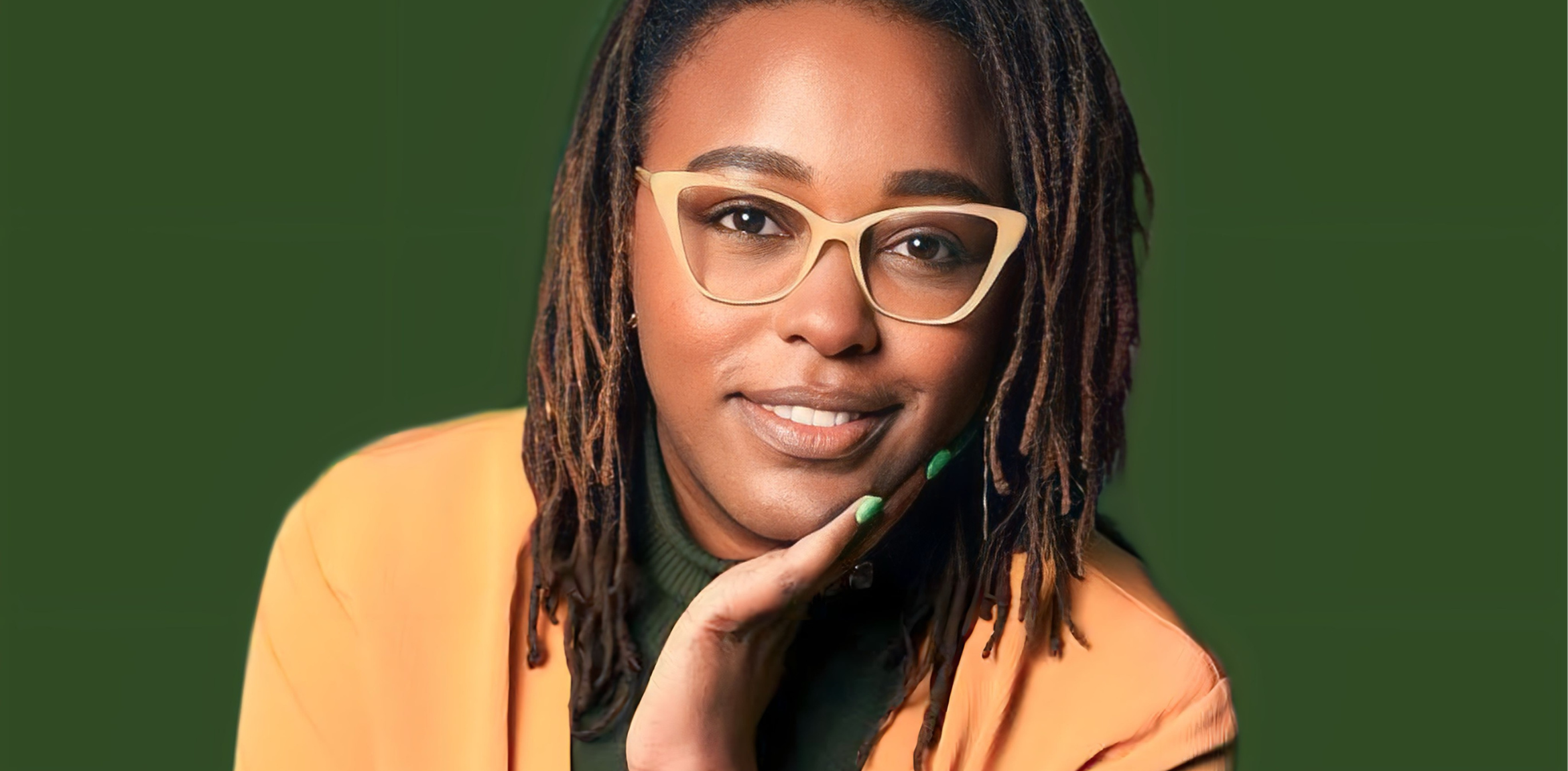 A professional woman with medium-length dreadlocks is shown in this image. She is wearing beige-framed glasses, a green top, and an orange blazer. She has a pleasant expression with a slight smile, resting her chin on her hand. The background is a solid green color.