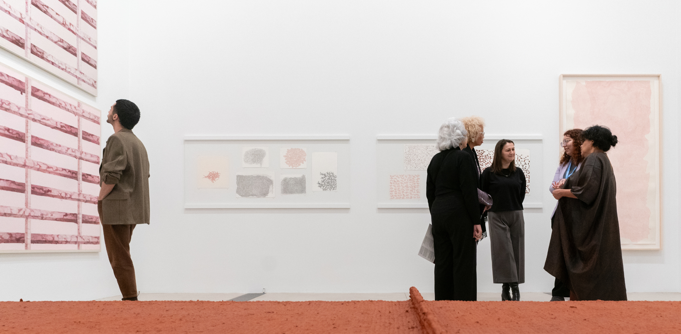 People in an art gallery viewing and discussing various abstract artworks on the walls. One man stands alone examining a large piece on the left, while a group of women are engaged in conversation near the center. The floor is covered with a textured, reddish-brown material, adding to the exhibition's aesthetic.
