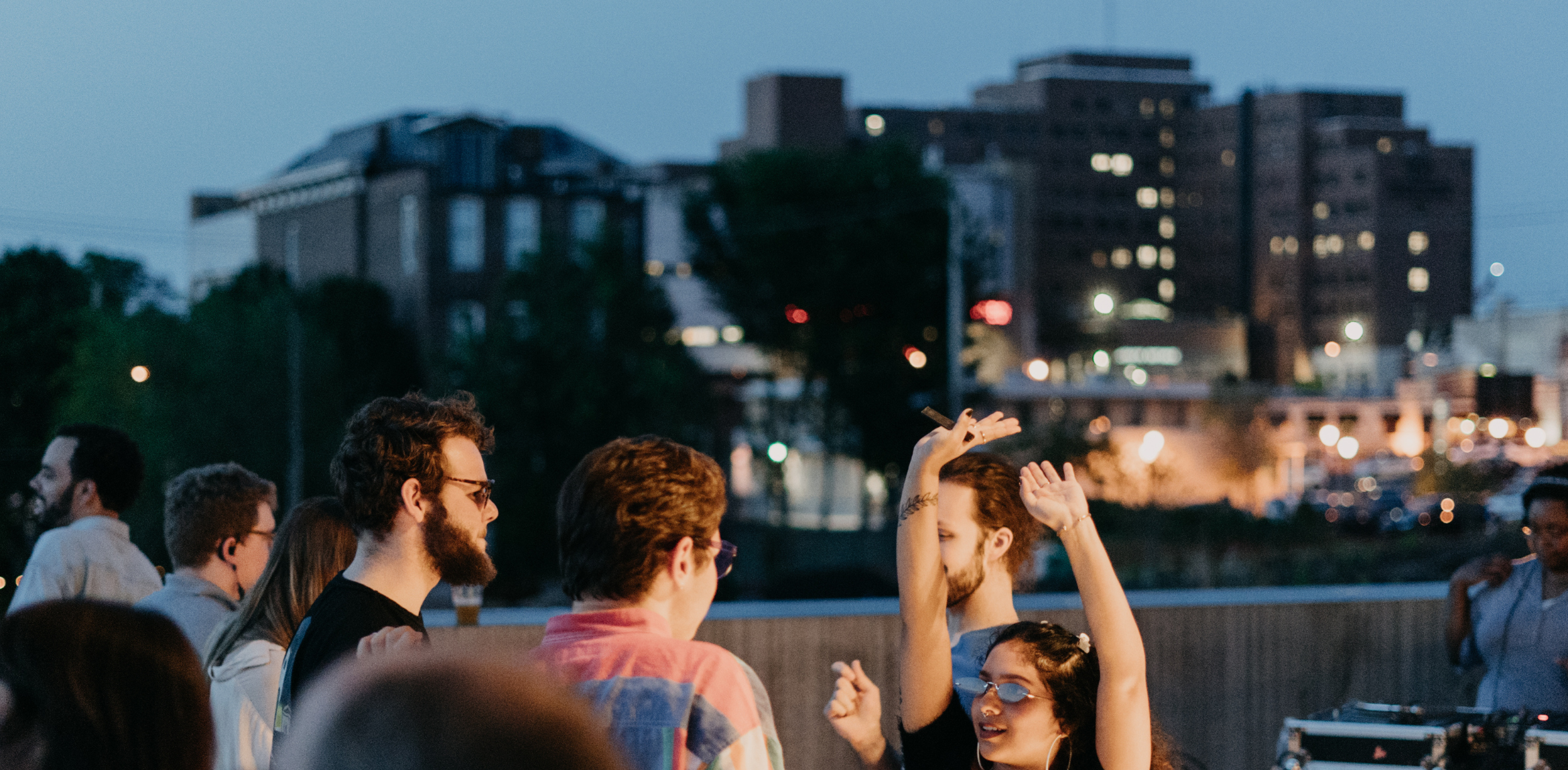 A group of people are dancing outdoors in the evening, with a cityscape in the background. Some individuals have their hands in the air, enjoying the moment. The scene includes a mix of people socializing and dancing, illuminated by the soft evening light. The background features buildings with lit windows, indicating a lively urban setting.