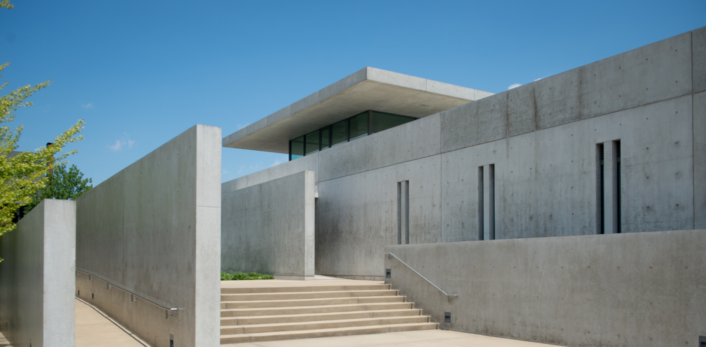 This image shows a modern, minimalist concrete building designed with clean lines and geometric shapes. The structure features a flat roof and narrow vertical windows. A set of stairs leads up to the main entrance, flanked by tall concrete walls. The building's aesthetic is characterized by the use of exposed concrete and an emphasis on simplicity and functionality. A small strip of greenery is visible on the left side, adding a touch of nature to the otherwise stark architectural design. The sky is clear and blue, providing a bright and clean backdrop for the building.