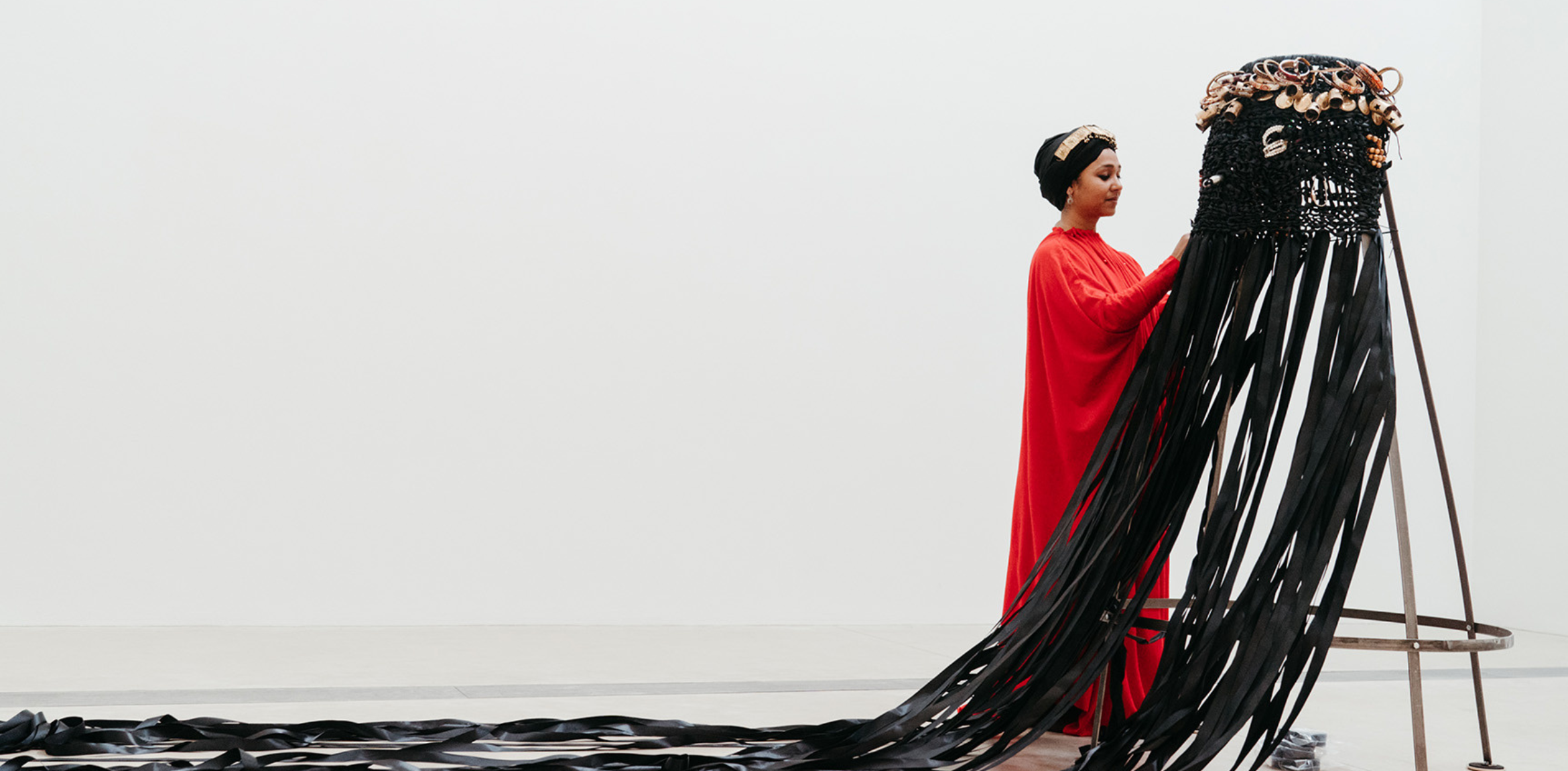 A person dressed in a long, flowing red garment and a black headwrap adorned with a gold headpiece is standing in an art gallery. They are interacting with a large, black, intricately woven sculpture that has long, black ribbon-like extensions flowing down from it and spread across the floor. The sculpture stands on a metal frame, and the background is a plain white wall, creating a minimalistic and modern setting.