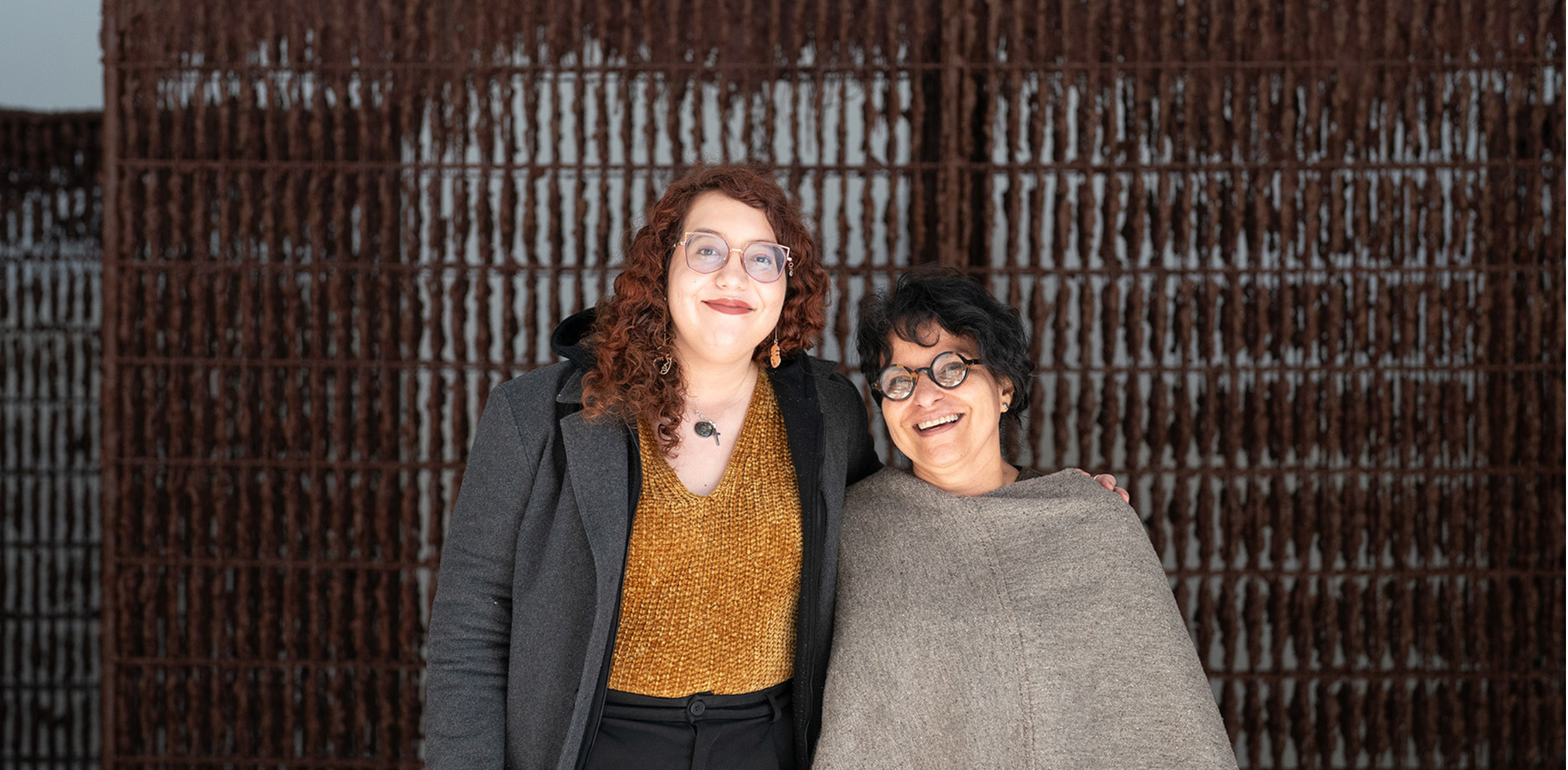 The image shows two smiling individuals standing close together against a background of a textured, vertical grid-like structure. The person on the left has curly red hair, wears glasses, and is dressed in a dark coat over a gold-colored top. The person on the right has short black hair, wears glasses, and is dressed in a beige wrap or shawl.