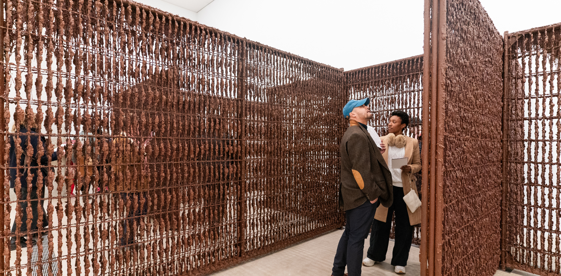 The image shows two people standing and interacting in front of a large, intricate structure made of what appears to be rusted metal bars with irregularly shaped material attached to them. The structure resembles a maze or enclosure with walls made of closely spaced vertical and horizontal bars, giving it a grid-like appearance. The two people, one wearing a blue cap and the other holding papers, seem to be engaged in conversation or examining the artwork.