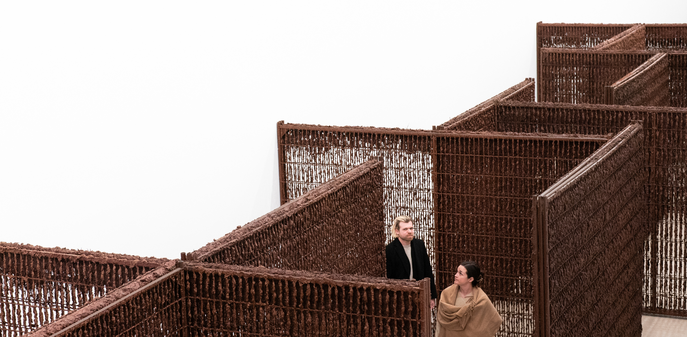 The image depicts two people standing in a maze-like structure made of rust-colored, woven metal or wire. The walls of the maze are tall and create a complex pattern of passages. The individuals appear to be observing their surroundings within the intricate layout of the maze.