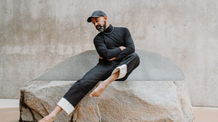 The image depicts a person sitting on a large rock, wearing a black turtleneck sweater, black pants with the cuffs rolled up to reveal the inner white lining, and a black cap. The person has a beard and is barefoot, with one leg extended out and the other folded underneath. The background is a simple, textured concrete wall. The pose suggests a sense of contemplation and balance.