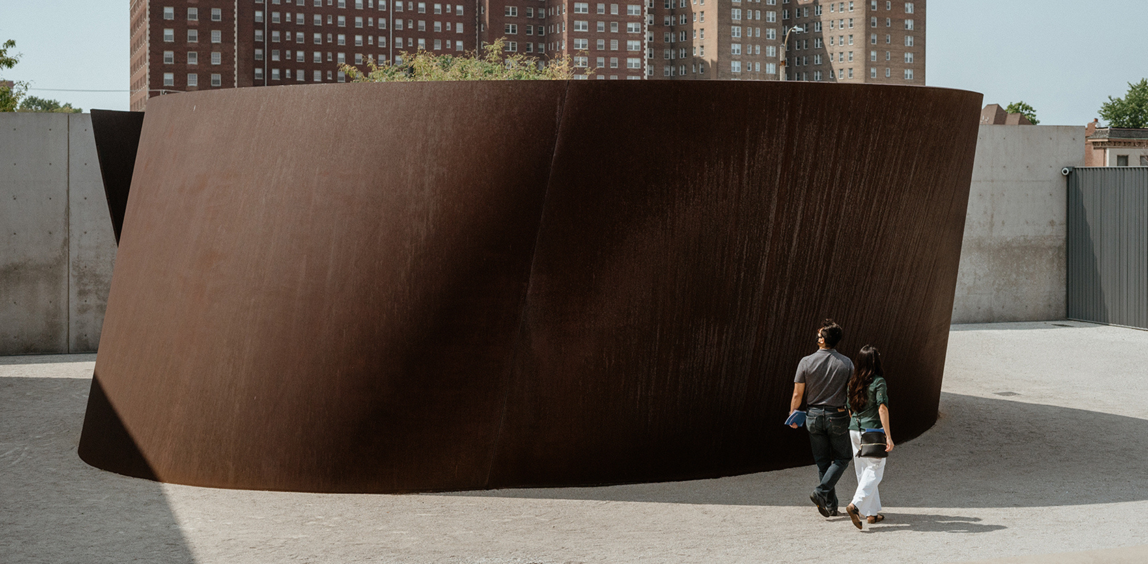 A couple walking past a large, curved, rust-colored sculpture in an outdoor space with a cityscape in the background.