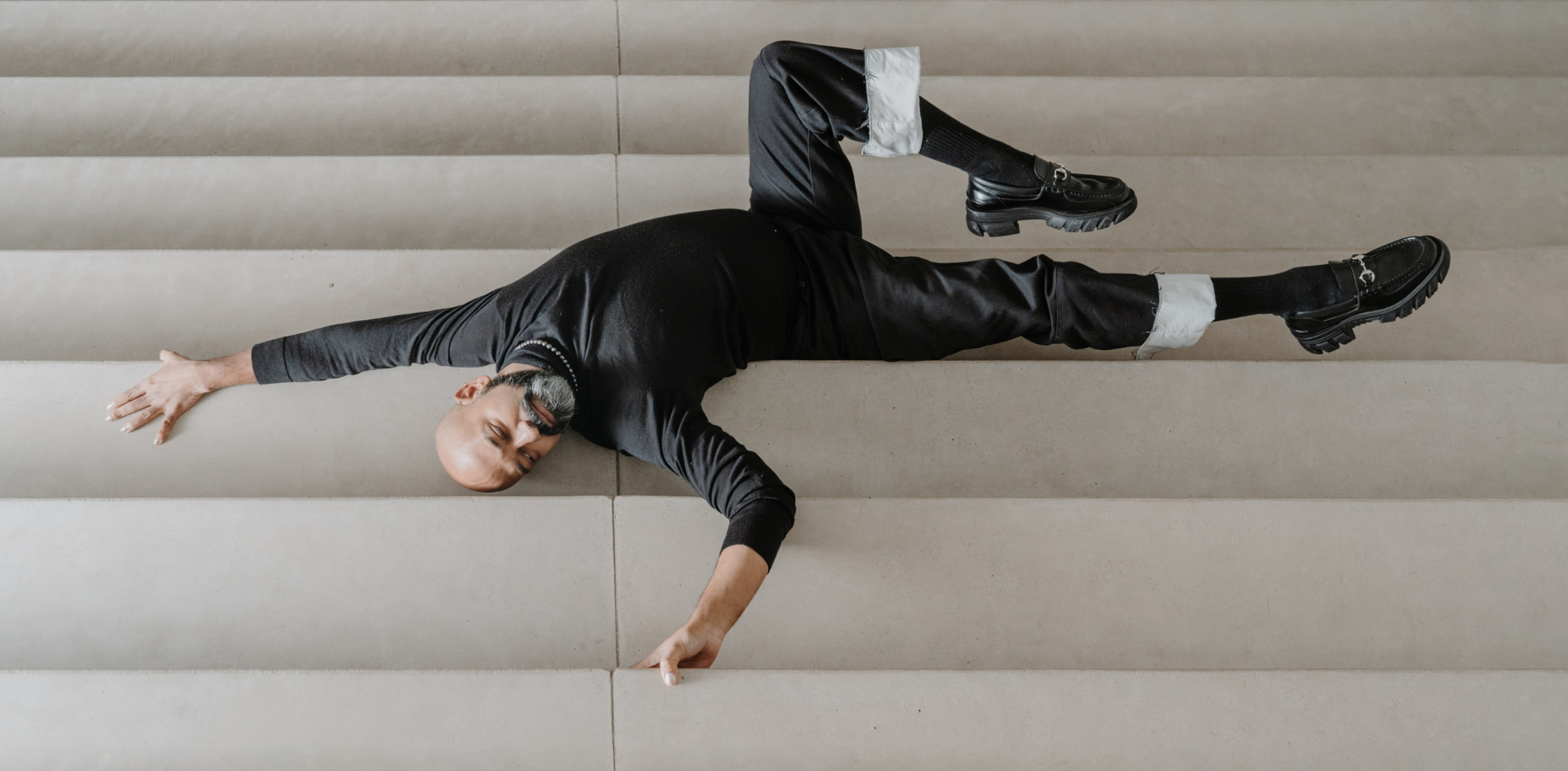 The image shows a man with a shaved head and beard lying in a striking pose on a set of light-colored, wide steps. He is dressed in all black with the cuffs of his pants rolled up, revealing white fabric. His arms and legs are extended diagonally in a dynamic position. He is wearing black loafers, and the setting appears minimalist with neutral tones, focusing on his body and posture against the backdrop of the stairs.