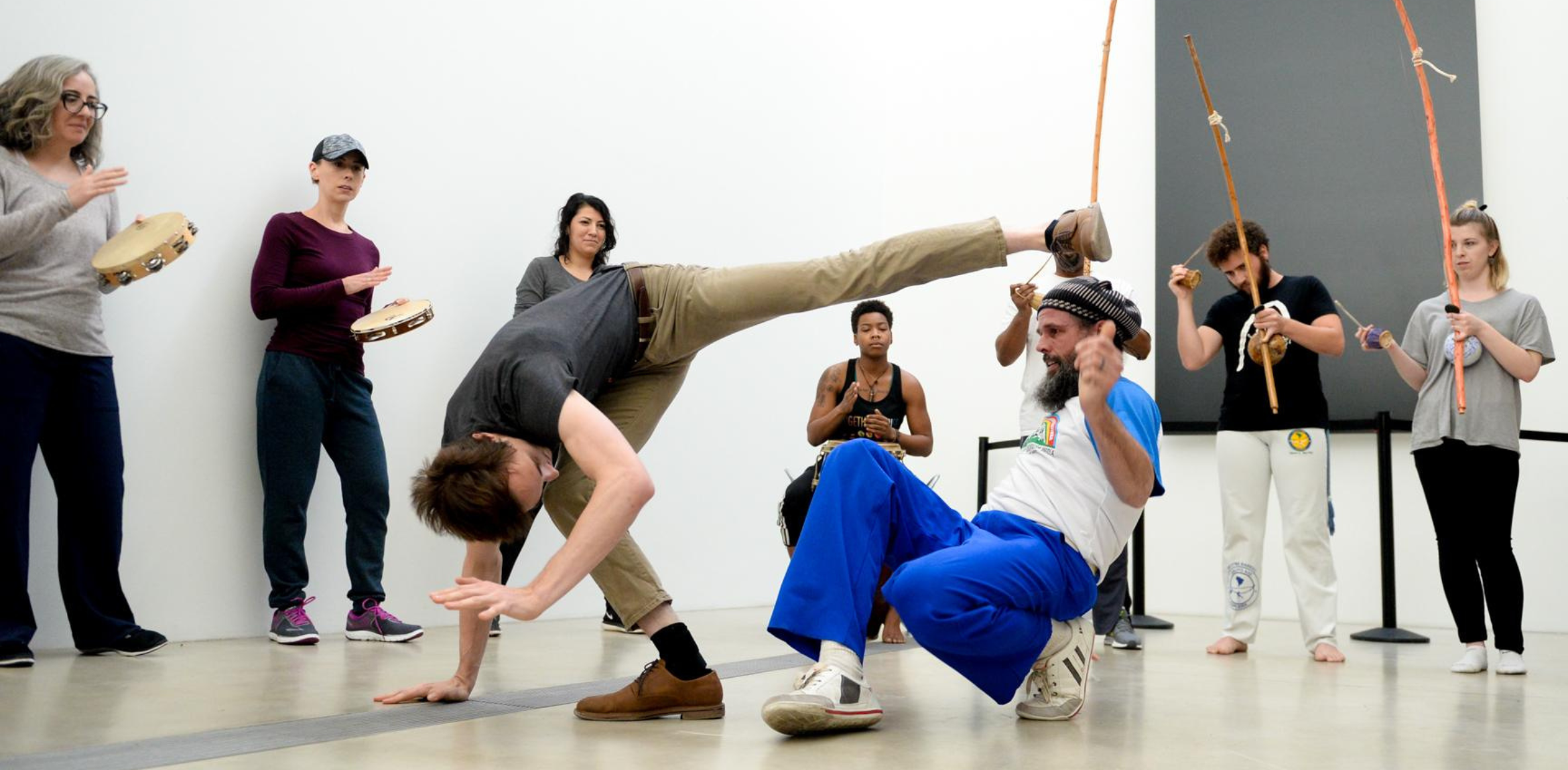 A group of people participating in a Capoeira session. In the foreground, two men are engaged in Capoeira movements: one is in mid-kick while balancing on one hand, and the other is crouching with one hand raised. Surrounding them, other participants are playing traditional Capoeira instruments, including tambourines and berimbaus. The scene takes place indoors against a plain white wall.