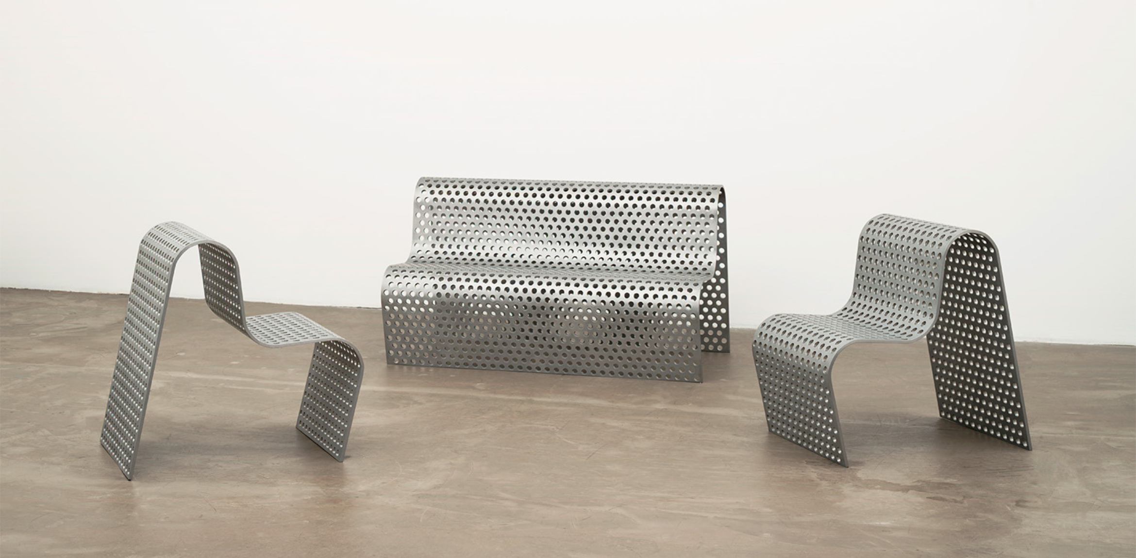 The image shows a set of furniture pieces designed by Scott Burton, consisting of a perforated metal settee and two perforated metal chairs. The pieces feature a sleek, modern design with a uniform pattern of circular holes throughout the metal surface, giving them a distinct, airy appearance. The settee has a curved backrest and seat, while the chairs have a continuous wave-like form that flows from the seat to the legs. The furniture is arranged in a minimalist setting with a smooth concrete floor and a plain white background, highlighting their sculptural quality.