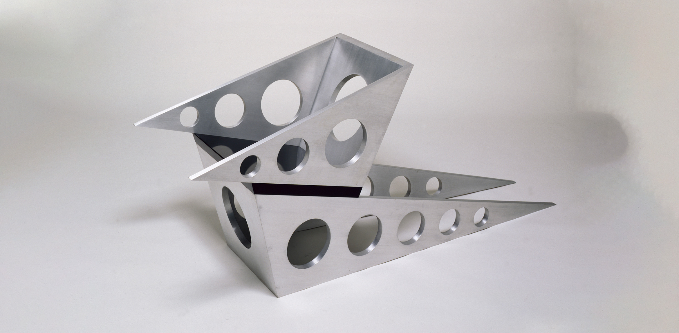 A metal sculpture with intersecting triangular shapes and circular cutouts, featuring a sleek, polished surface. The piece has a geometric and modern design.