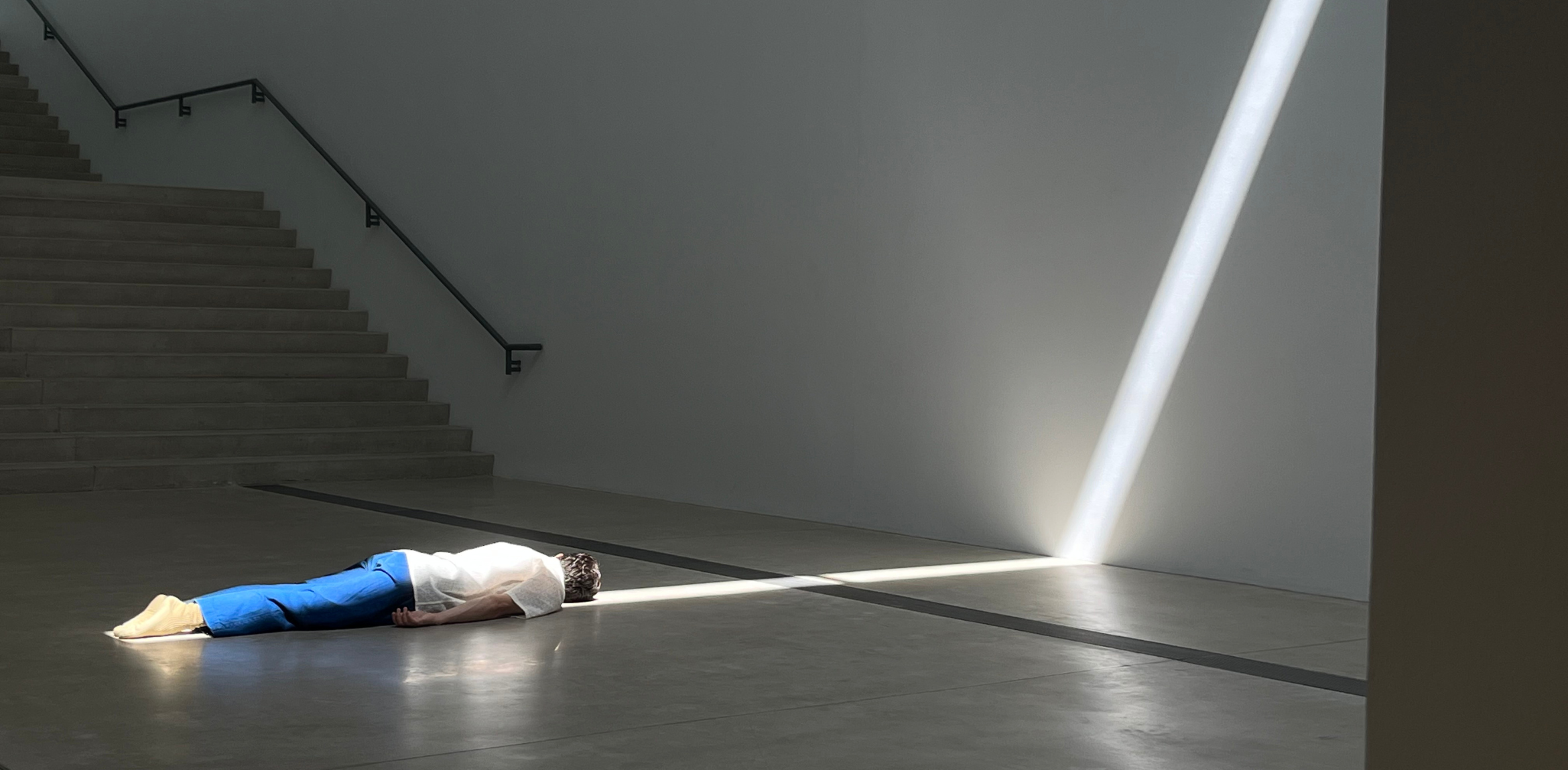 The image shows a person lying on the floor, partially illuminated by a beam of light coming through a narrow opening on the right side. The individual is wearing a light-colored top and blue pants, with their head and upper body in the beam of light, while their lower body remains in shadow. The surrounding space appears minimalist, with a staircase on the left side and smooth, clean walls. The contrast between the bright light and the darker surroundings creates a dramatic and contemplative atmosphere.