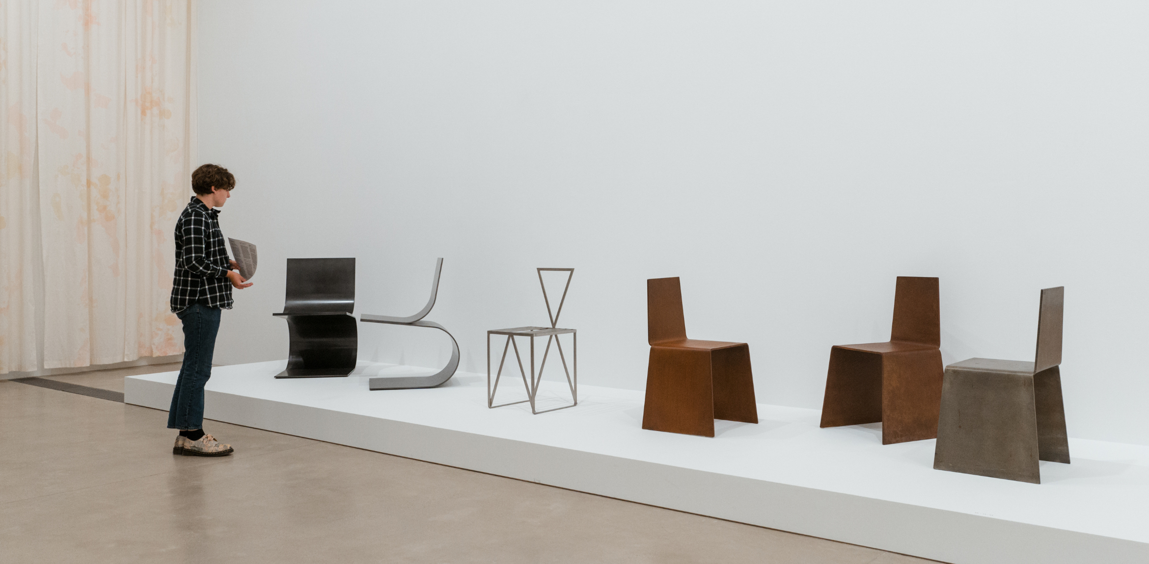 The image depicts a person standing in front of a display of modern, minimalist chairs. The person is wearing a checkered shirt, cropped jeans, and patterned shoes, holding a piece of paper, possibly observing the exhibit. The chairs are arranged in a row on a raised white platform against a plain white wall. Each chair is distinct, with geometric and industrial designs—some made of metal and others with rust-like finishes. The scene takes place in what appears to be a gallery or museum with light flooring and soft, neutral-colored curtains hanging in the background.