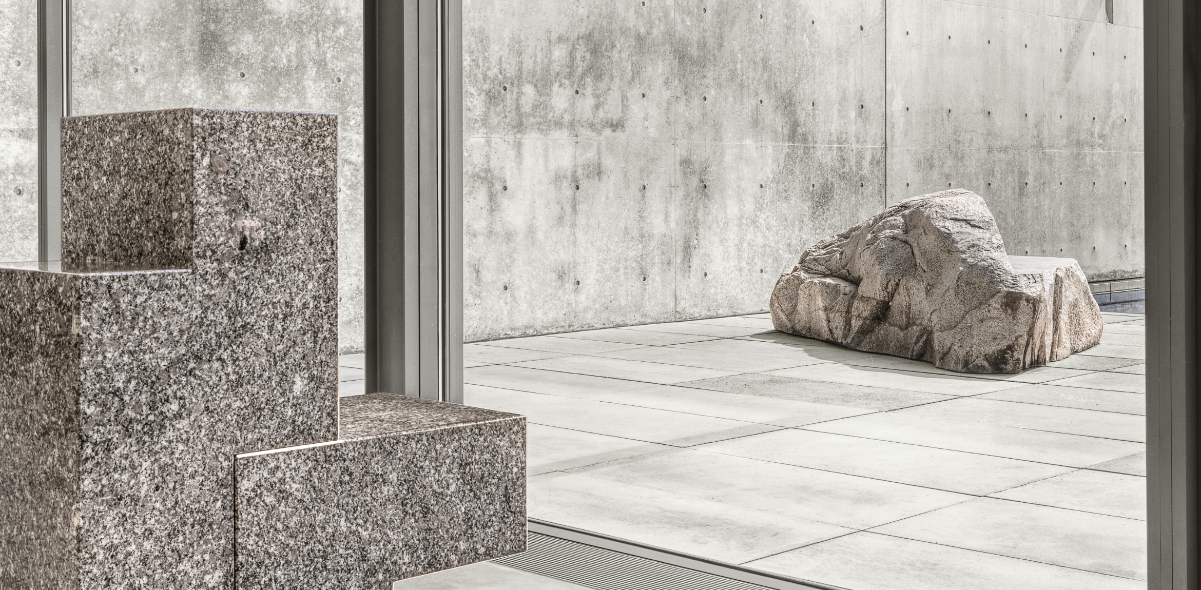 The image shows two stone sculptures in a modern architectural setting. In the foreground, there's a polished, geometric stone sculpture with a rectangular, blocky shape, consisting of stacked square forms. This sculpture is positioned indoors, near a glass window. In the background, visible through the window, there is a large, natural rock formation placed outdoors on a smooth stone surface. The setting is minimalist, with the outdoor area framed by clean concrete walls, which have a textured, weathered look, enhancing the contrast between the natural and geometric forms of the sculptures.