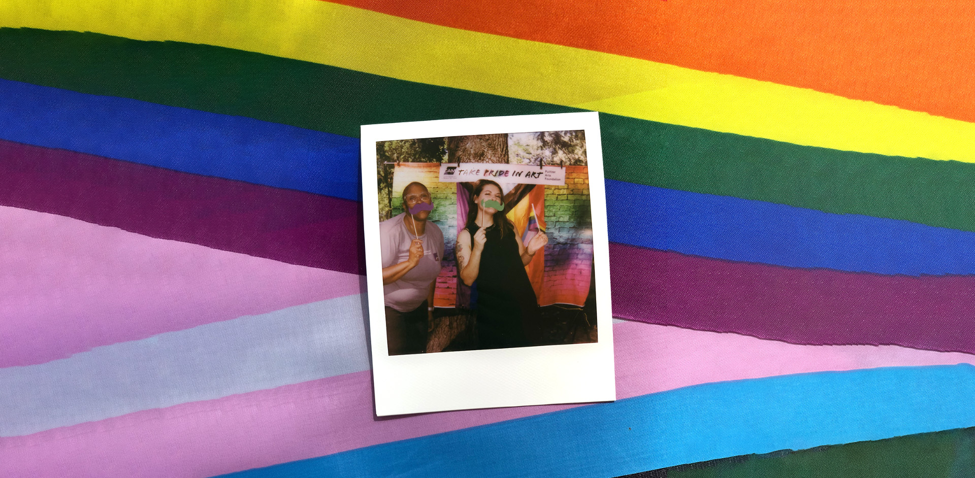 The image features a polaroid-style photo of two individuals holding colorful mustache props, standing in front of a vibrant backdrop. The backdrop displays the message 