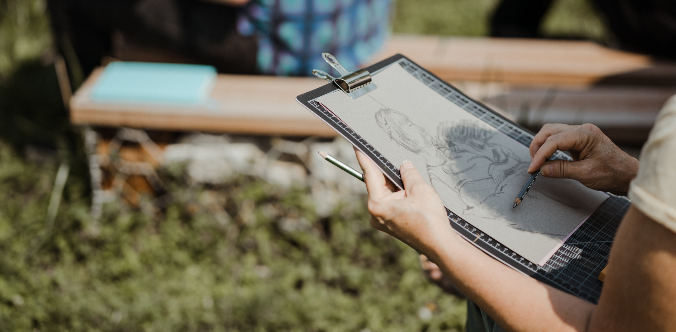 This image shows a person sketching outdoors on a clipboard. The drawing in progress appears to be a human figure. The person's hands are holding a pencil and another sketching tool, and they are seated on a bench in the background, which seems to be surrounded by greenery. The focus is on the hands, sketchbook, and drawing, with the surroundings slightly blurred.