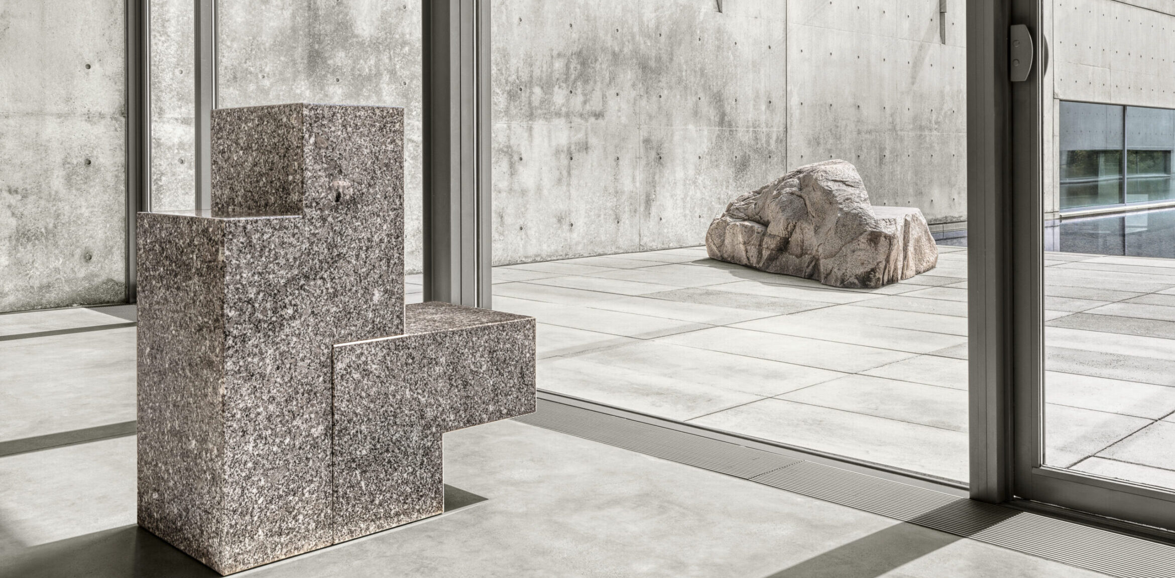 Image of a granite chair in front of a window. A rock sculpture is in the background surrounded by concrete.