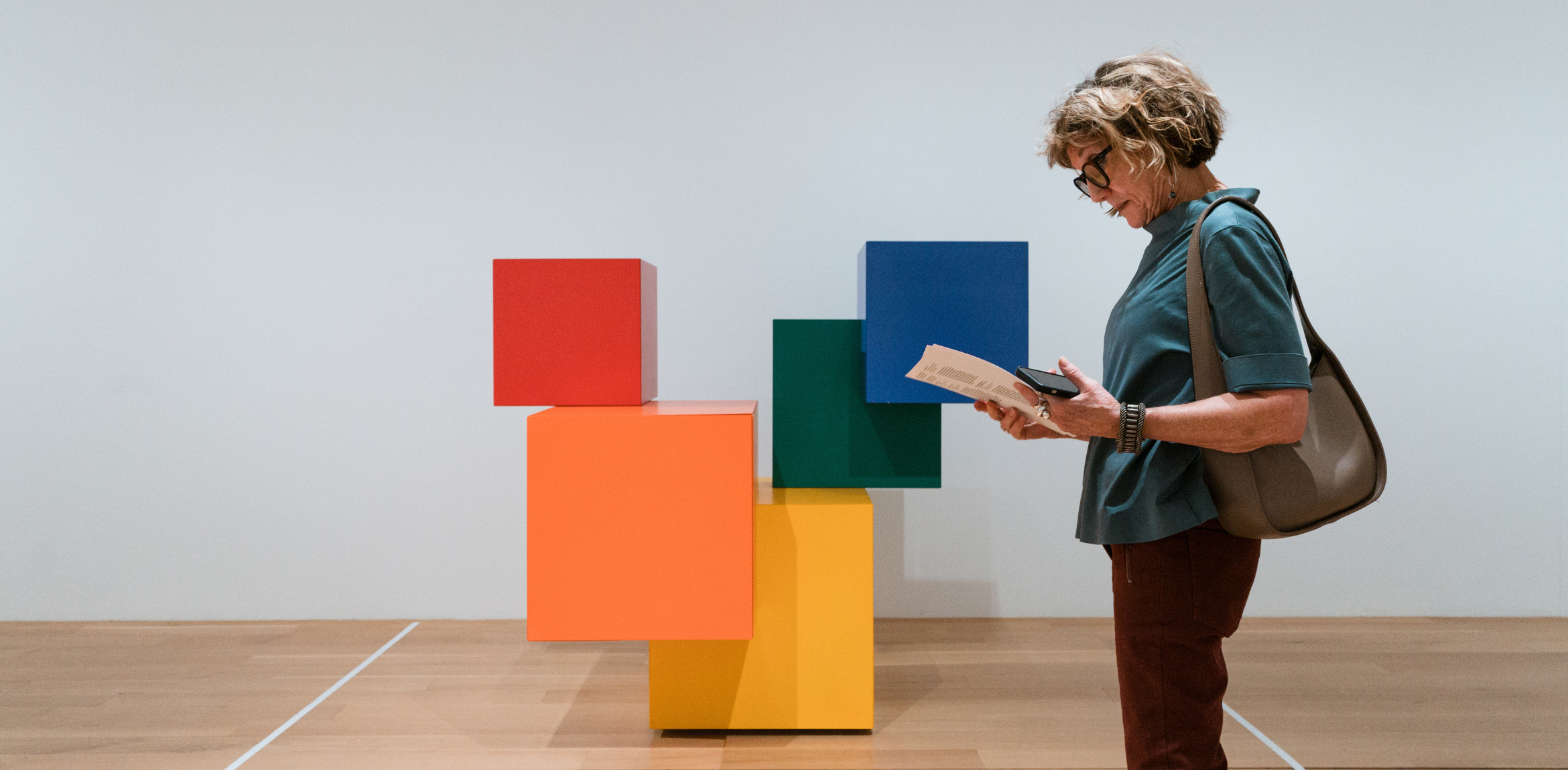 The image shows a person standing indoors, holding a document or brochure while examining an art installation. The artwork features five brightly colored cubes, stacked in an abstract arrangement, with red, orange, yellow, green, and blue blocks forming a geometric structure. The cubes are positioned on a wooden floor, and the background is a plain white wall. The person in the image is dressed casually in a teal top, maroon pants, and glasses, with a taupe-colored shoulder bag, focusing intently on what they are reading.