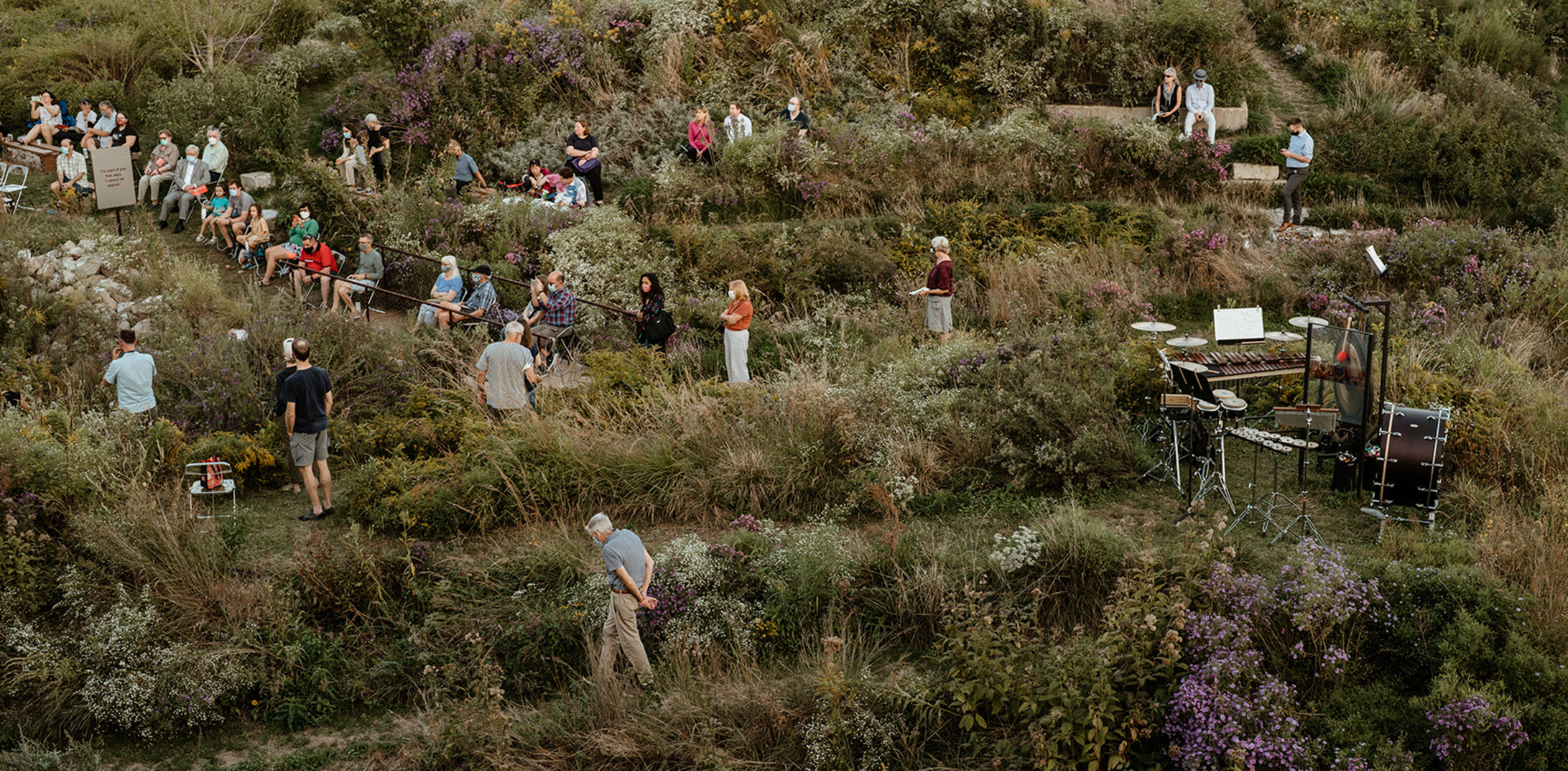 The image shows a natural outdoor setting with people seated and standing among wild grasses, bushes, and purple flowers. The attendees appear to be scattered across various levels of terrain, which resembles a small amphitheater or hillside garden. Some people sit on benches, while others stand or walk. A percussion setup with various instruments is placed on the right side, ready for a performance. The atmosphere is serene, blending art and nature, with the audience integrating into the landscape.